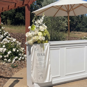 May include: A white bar with a floral arrangement and a white parasol. A white banner reads "the BAR" with drink options. The floral arrangement includes white and green flowers. The bar is outdoors with a natural background.