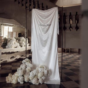 May include: A white fabric seating plan with the text "Your Seat Awaits" and guest names, hanging from a dark metal frame. White floral arrangements are at the base. The setting is a formal event space.