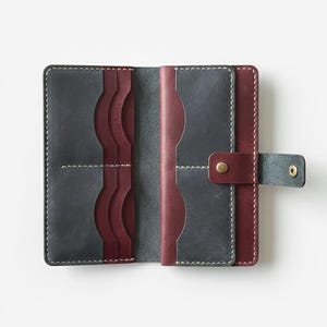 May include: Open leather wallet in gray and burgundy. The wallet has multiple card slots and a snap closure. White stitching accents the edges. The interior is burgundy, and the exterior is gray. The wallet is open, displaying its interior.