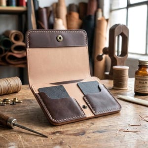 May include: Open leather wallet with brown and tan tones, featuring card slots and a snap closure. The wallet is handcrafted with visible stitching and sits on a wooden surface, showcasing the craftsmanship.