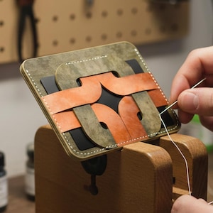 Könnte beinhalten: Eine handgefertigte Ledergeldbörse in Grün-, Orange- und Schwarztönen wird genäht. Die Geldbörse hat ein einzigartiges Webdesign und wird in einem hölzernen Nähpferd gehalten. Der Hintergrund zeigt Werkzeuge und Materialien in einer Werkstatteinstellung.