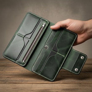 Könnte beinhalten: Offene, grüne Ledergeldbörse mit mehreren Fächern, einer Reißverschlusstasche und einem Druckknopfverschluss. Die Geldbörse ist mit weißem Faden genäht und hat ein abgenutztes, strukturiertes Aussehen. Das Innere ist sichtbar.