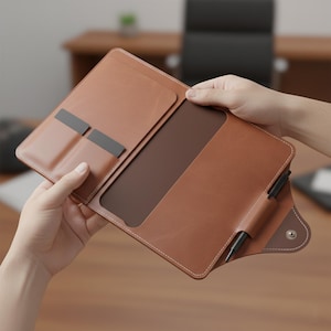 May include: A tan leather portfolio, open to show a dark brown interior and card slots. White stitching outlines the edges. A black pen is held in a loop. The portfolio is held open.