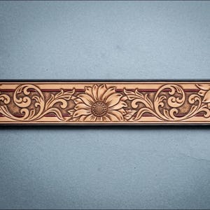 Op de afbeelding: Close-up van een bruinleren riem met een gebeeldhouwd zonnebloem- en krulontwerp. De riem heeft een donkerbruine rand en een rode streep. De zonnebloem staat in het midden, met gedetailleerde bloemblaadjes en een getextureerd centrum.