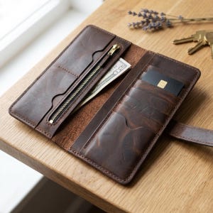 May include: Open brown leather wallet with multiple card slots, a zippered pocket, and a bill compartment. The wallet is open, revealing a credit card and a US $100 bill. The wallet is on a wooden surface.