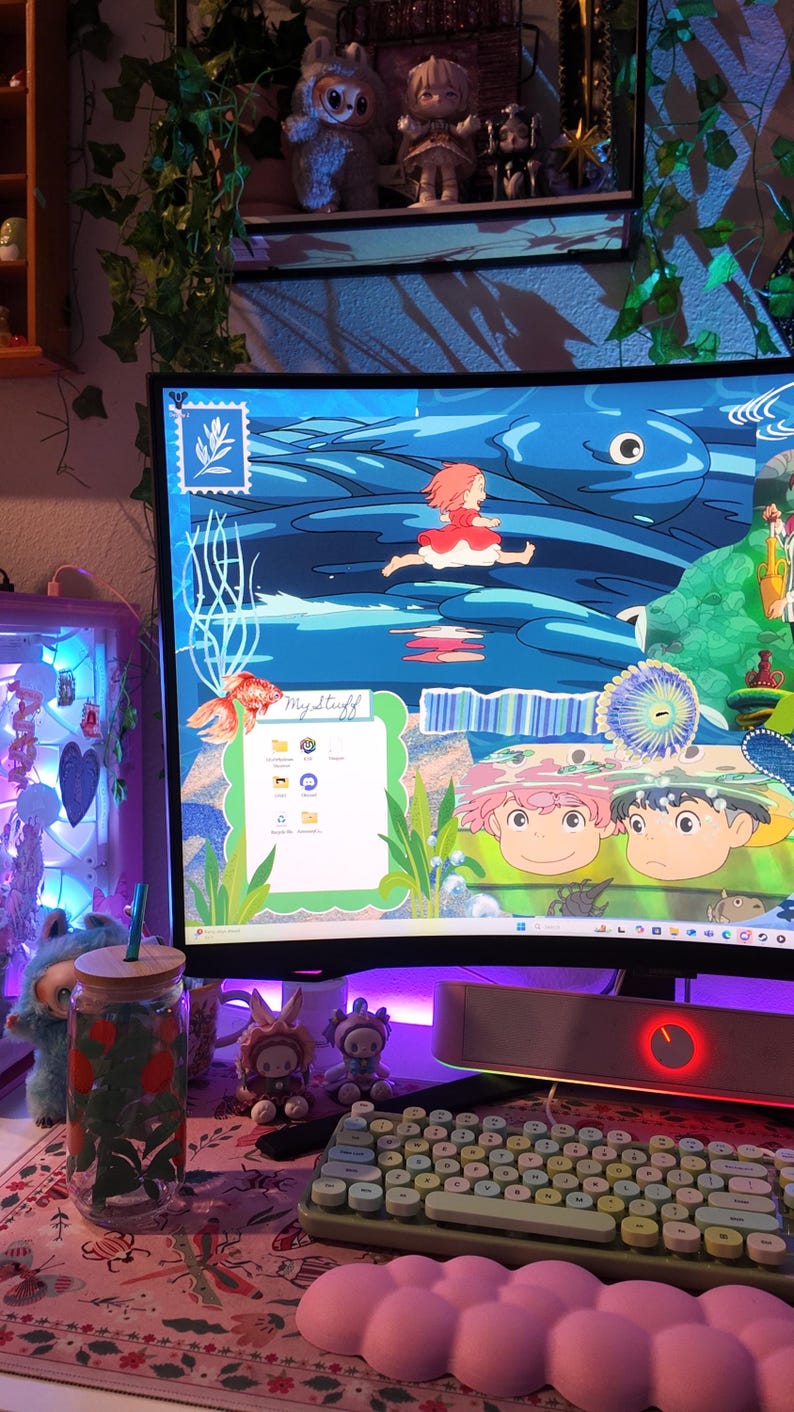 May include: A desktop computer with a colorful animated wallpaper featuring a girl in a red dress running across a blue ocean. A green and white keyboard, a pink cloud-shaped wrist rest, and a glass jar with a straw are in the foreground.
