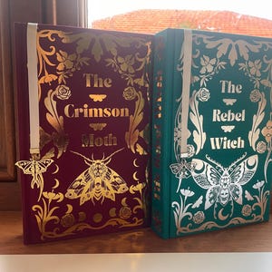 Puede incluir: Dos libros de tapa dura con cubiertas decorativas. Uno es burdeos con detalles dorados y el título "The Crimson Moth". El otro es verde azulado con detalles plateados y el título "The Rebel Witch". Ambos tienen marcapáginas con forma de mariposa.