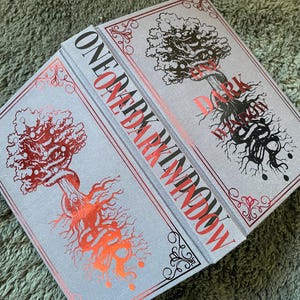 Puede incluir: Se muestran dos libros de tapa dura titulados "One Dark Window". Los libros tienen una cubierta gris claro con detalles en rojo y negro. La portada presenta un diseño de árbol con detalles intrincados y el texto del título en rojo.