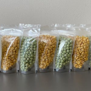 May include: A series of transparent plastic bags, each containing dried peas or corn kernels. The bags alternate between green peas and yellow corn, creating a colourful display. The bags are sealed and stand upright.