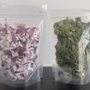 May include: Two clear plastic bags filled with dried vegetables. One bag contains chopped red onions, and the other contains dried green onions. The bags are sealed and ready for use.