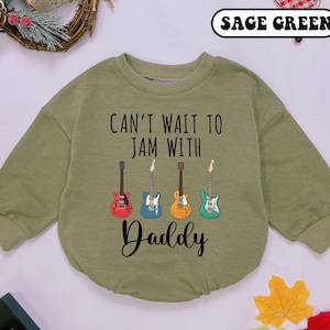 May include: Sage green baby romper with long sleeves. The romper features the text "Can't wait to jam with Daddy" and four colorful guitar illustrations. A cute and comfortable outfit for a little musician.