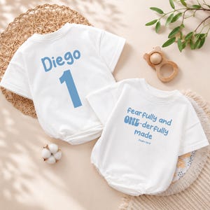May include: Two white baby onesies. One has the name "Diego" and the number "1" in blue. The other onesie has the text "fearfully and ONE-derfully made" in blue. A wooden toy and cotton ball are also visible.