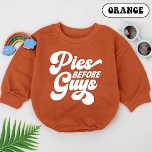 May include: An orange baby romper with the text "Pies Before Guys" in white. The long-sleeved romper has a round neckline and is paired with light blue sunglasses and a rainbow decoration.