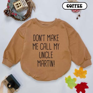 May include: A coffee-colored baby sweatshirt with the text "DON'T MAKE ME CALL MY UNCLE MARTIN!" in black. The long-sleeved top has a round neckline and a relaxed fit. The image includes decorative fall leaves and a small Christmas decoration.