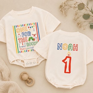 May include: Two cream-colored short-sleeve bodysuits. One features a colorful graphic with the text "ONE FOR THE BOOKS" and "Noah's first birthday" with a caterpillar and cake. The other features the name "NOAH" above a large red number one candle.