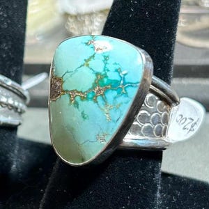 May include: A silver ring featuring a large, triangular, light blue turquoise stone with dark brown matrix veining. The band is crafted from polished silver and displays a textured, repeating scale-like pattern along the sides of the metal shank.