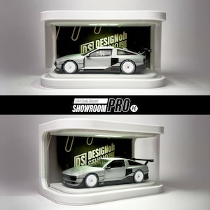 May include: A silver diecast model car displayed in a white case. The car features a black spoiler and white wheels with red centres. The display case has the text "DS DESIGNoh" and "SHOWROOM PRO".
