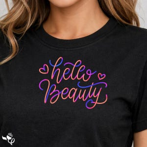 May include: A black t-shirt featuring the text "hello Beauty" in a cursive, multi-coloured gradient font that transitions between shades of pink, orange, and purple. Two small hearts are positioned on either side of the word hello.
