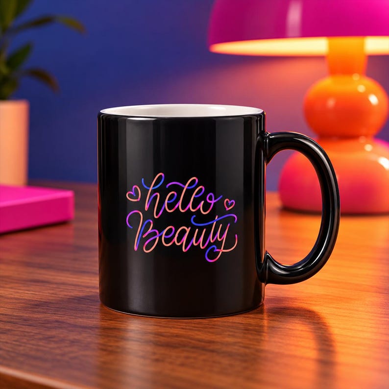 May include: A black ceramic mug with a white interior and a handle on the right side. The front of the mug features the text "hello Beauty" written in a stylised, cursive font with a gradient of pink, purple, and orange colours, accented by small hearts.