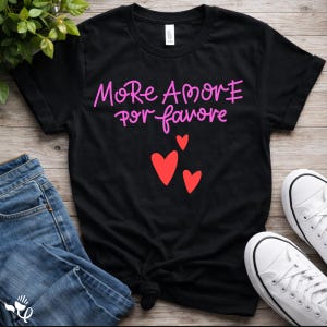 May include: A black short-sleeved t-shirt featuring the pink text More Amore por favore printed on the chest. Below the text are three red heart shapes of varying sizes. The shirt has a crew neckline and a standard fit, displayed flat.
