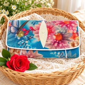 May include: A rectangular clutch bag featuring a vibrant floral and butterfly print in shades of blue, pink, and orange. The surface has a textured, pebbled finish with a central, elongated, cream-coloured oval accent piece on the front flap.