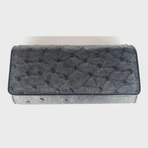 May include: A dark grey clutch wallet with a textured ostrich leather design. The wallet has a rectangular shape with rounded edges and a flap closure. The surface is covered in small, raised bumps, and the edges are stitched.