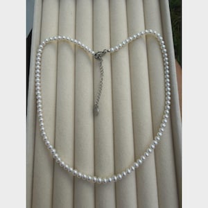 Può includere: Una delicata collana di perle con chiusura argentata e catena di estensione. La collana presenta piccole perle bianche rotonde infilate insieme. La collana è esposta su una superficie beige chiaro.