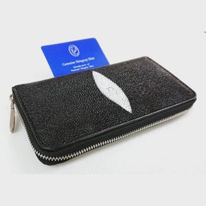 Könnte beinhalten: Schwarzes Portemonnaie aus Stachelrochenhaut mit einem weißen, augenförmigen Design. Die Geldbörse hat einen silbernen Reißverschluss und eine blaue Karte mit der Aufschrift "Genuine Stingray Skin". Die Geldbörse ist rechteckig und scheint neu zu sein.