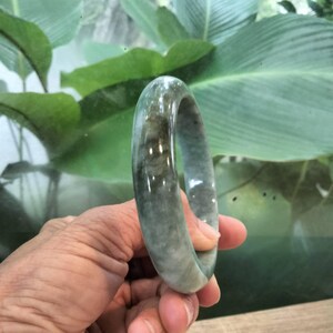 以下が含まれることがあります： 緑と灰色の斑点模様がある石で作られた、滑らかで磨き上げられたバングルブレスレット。円形のアクセサリーは丸みを帯びた形状で、自然な色の変化があり、全体的に半透明の質感と光沢のある仕上げが施されています。