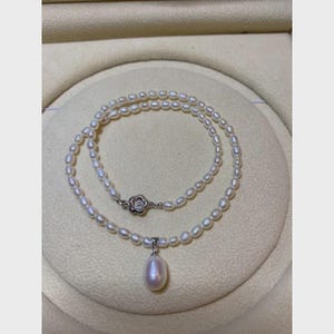 Può includere: Una delicata collana di perle con un design a doppio filo. La collana presenta piccole perle rotonde e un pendente a goccia più grande. La chiusura è color argento con un motivo floreale. La collana è esposta su una superficie color crema.