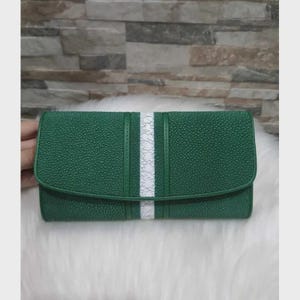 May include: A green wallet with a textured surface and a white stripe down the center. The wallet is rectangular with a curved flap closure. The wallet is resting on a white, fluffy surface.