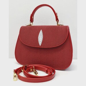 May include: A red handbag with a top handle and a detachable shoulder strap. The bag features a textured surface and a white, oval-shaped design on the front. Gold-colored hardware accents the bag and strap.
