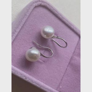 Peut inclure: Une paire de boucles d'oreilles en perles blanches avec un design en crochet argenté. Les perles sont rondes et ont une finition brillante. Les boucles d'oreilles sont présentées dans un écrin en velours violet.