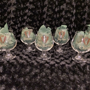 May include: A set of seven clear brandy snifters, each personalized with a name and a tuxedo graphic. The glasses are filled with green tissue paper. The names include Dra, Jroc, James, Jay-lan, Chris, and Davi.