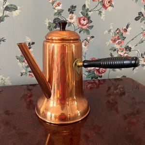 May include: A polished copper coffee pot featuring a tall cylindrical body, a long angled spout, and a black wooden side handle. The pot includes a matching copper lid topped with a small black knob, showcasing a reflective metallic finish.