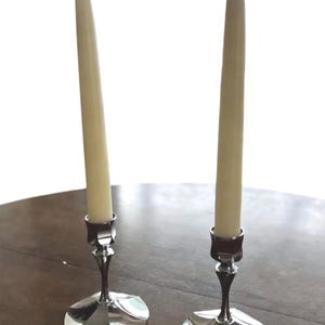 May include: Two silver candle holders with cream-colored candles. The candle holders have a decorative base and a slender stem. The candles are tall and slender, with a curved top. The items are on a dark wooden surface.