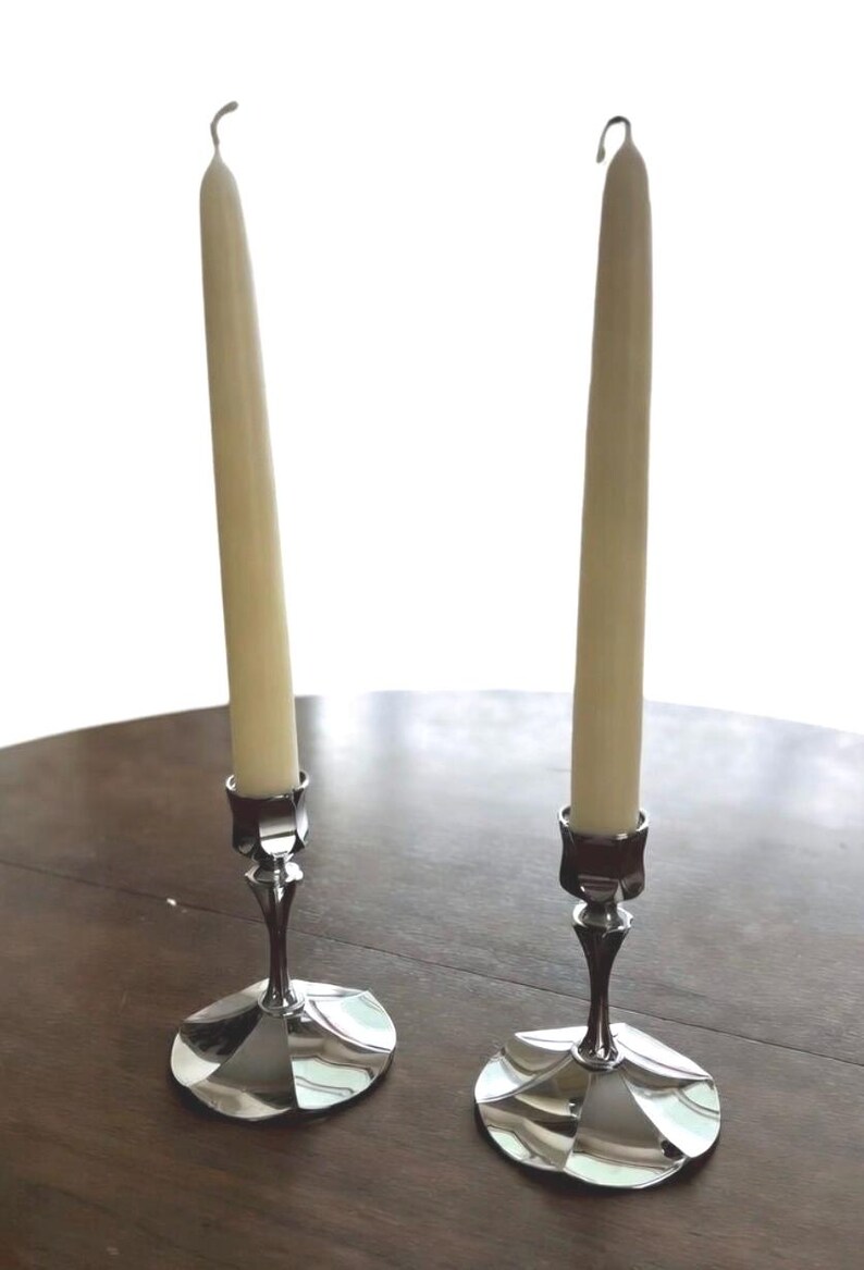 May include: Two silver candle holders with cream-colored candles. The candle holders have a decorative base and a slender stem. The candles are tall and slender, with a curved top. The items are on a dark wooden surface.