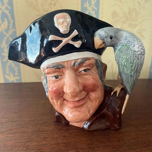 May include: A ceramic pirate figurine with a black hat featuring a skull and crossbones, and a parrot perched on the shoulder. The pirate has a smiling face and is wearing a brown coat. The parrot is gray and green.