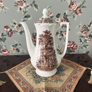 May include: A white ceramic coffee pot featuring a brown transferware print of a colonial-style building with a clock tower and figures in the foreground. The pot has a tall, slender spout, a curved handle, and a matching lid with a finial top.