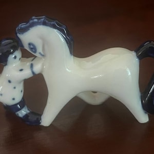 May include: A white ceramic figurine featuring a stylized horse with a dark blue mane and tail, standing beside a small figure with dark blue hair and a white shirt with blue polka dots, both connected in a single smooth, glossy porcelain sculpture.