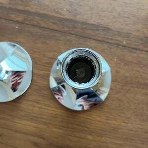 May include: Close-up of two shiny, silver bottle stoppers. The stoppers have a faceted design on the top and a cylindrical base. The stoppers are on a wooden surface.