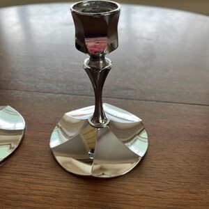 May include: A silver-colored candlestick with a geometric design. The candlestick has a wide, circular base and a slender stem that leads to a cup-shaped holder. The surface reflects the surrounding environment.