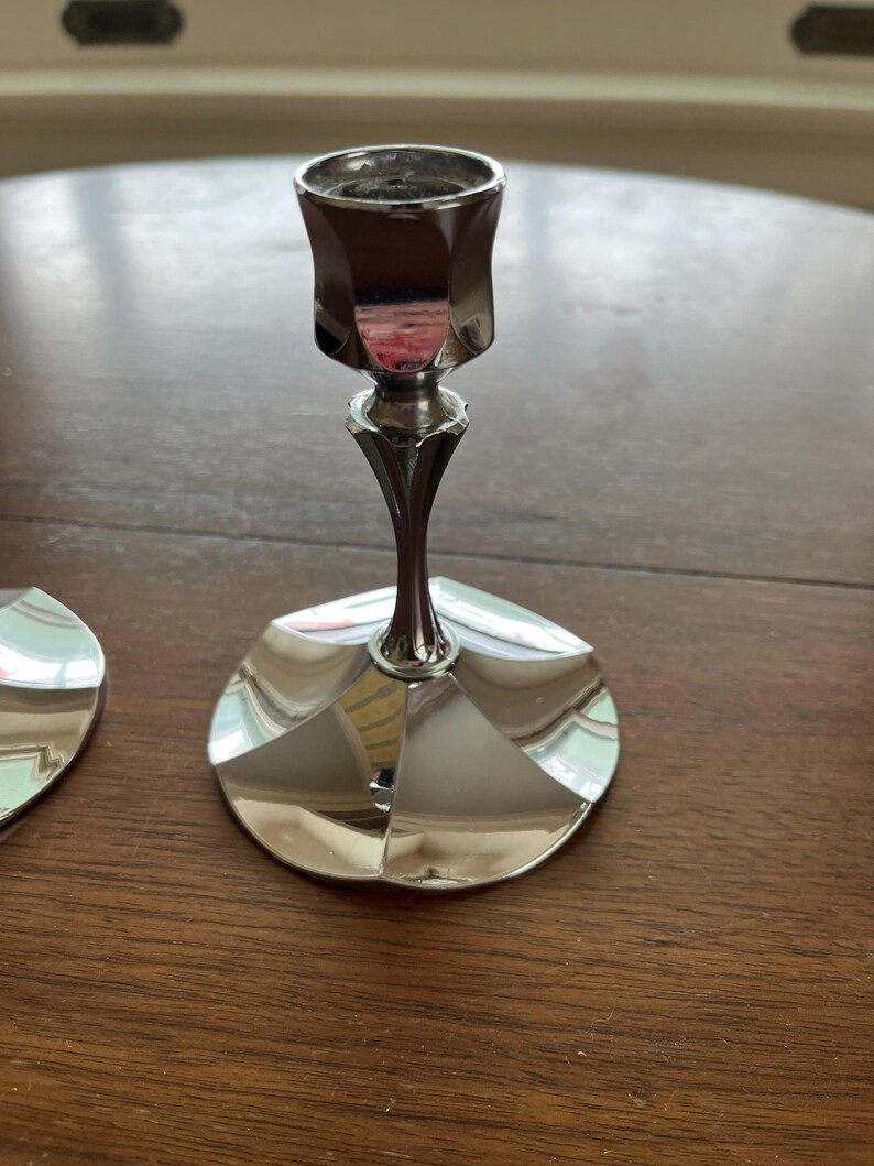 May include: A silver-colored candlestick with a geometric design. The candlestick has a wide, circular base and a slender stem that leads to a cup-shaped holder. The surface reflects the surrounding environment.