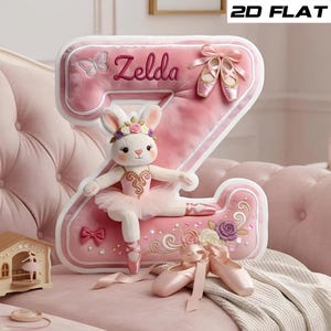 May include: A plush pink letter "Z" with the name "Zelda" embroidered on it, decorated with a ballerina bunny, ballet shoes, and floral accents. The bunny wears a tutu and flower crown. A small wooden ballerina music box is in the foreground.