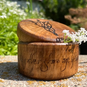 Könnte beinhalten: Runde Holzbox mit Deckel, mit geschnitztem Design und graviertem Text. Die Box ist geöffnet und gibt den Blick ins Innere frei, begleitet von weißen Blumen. Der Text auf der Box lautet "rather share one lifetime".