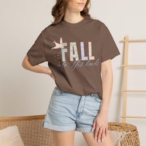 May include: A brown short-sleeved t-shirt featuring a graphic design with the word FALL in block letters filled with floral patterns, a small bird illustration, and the cursive text into His hands printed in a light blue color on the front.