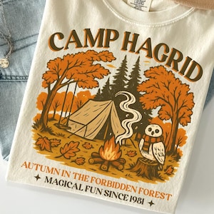 May include: Ivory-colored t-shirt with the text "CAMP HAGRID" and a graphic of a campsite with trees, a tent, and an owl. The shirt is paired with light blue jeans and a tan hat. The text "AUTUMN IN THE FORBIDDEN FOREST + MAGICAL FUN SINCE 1981 +" is also visible.