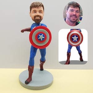 May include: A Captain America bobblehead figurine. The figure is wearing a blue suit, brown boots, and a red, white, and blue shield. The head is a likeness of a man with brown hair and a beard. The figurine is on a gray circular base.