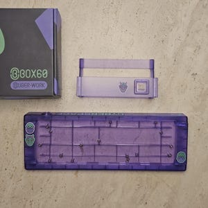 Peut inclure: Un boîtier de clavier violet et ses composants sont présentés sur une surface de couleur claire. Le boîtier est rectangulaire avec un motif de grille et de petites broches métalliques. La boîte indique "BOX60" et "@UGER-WORK".