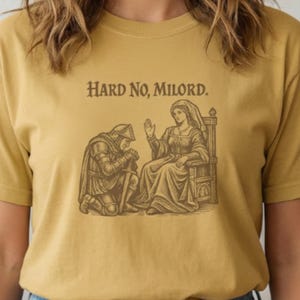 Può includere: T-shirt giallo senape con grafica in stile medievale e la scritta "Hard No, Milord." La grafica raffigura un cavaliere in ginocchio e una donna seduta. La maglietta è in materiale morbido.